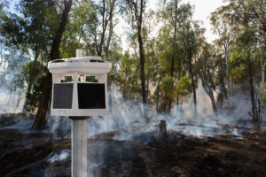 white all-in-one fire weather station stands on a pole mount in front of a controlled fire burn in a forest near Whitfield, King Valley, Victoria, Australia