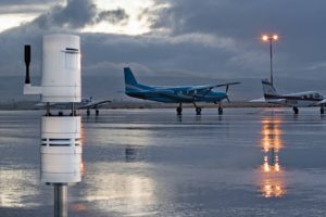 white automated weather observation system stands in front of high-wing airplane on a rainy runway at dusk