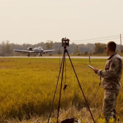 Soldier looks at data from weather station on a tablet in the foreground as a fighter jet takes off from a public highway in the background