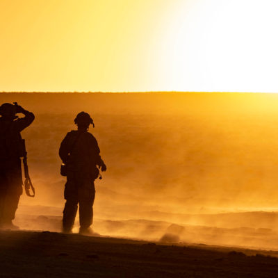 two soldiers stand in desert environment with sun on horizon creating a large orange glow