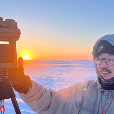 Man wearing glasses, a knit cap, and winter coat adjusts a micro weather station standing atop a tripod as the sun rises over a frozen tundra