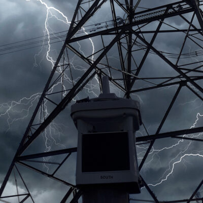 micro weather station stands in front of transmission tower amidst rain and lightning