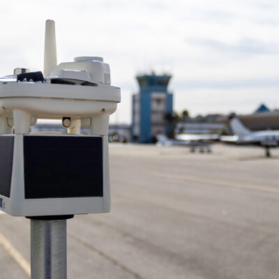 small white weather station stands in front of ga aircraft and air traffic control tower