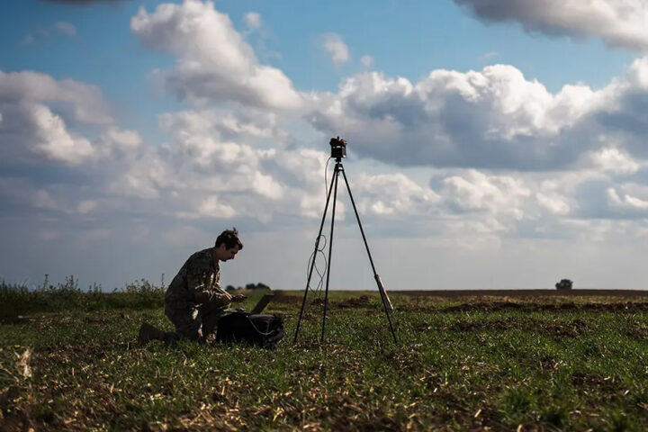 brown weather station on tripod with soldier in green fatigues on laptop in field