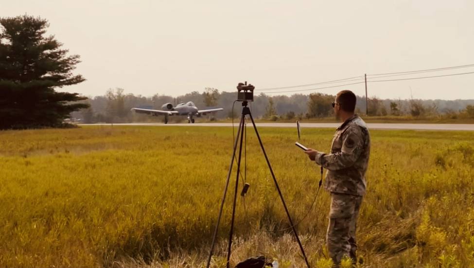 Soldier looks at data from weather station on a tablet in the foreground as a fighter jet takes off from a public highway in the background